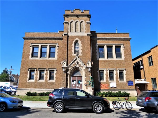 A tour bike between two cars in front of a brown brick two story former church and school with a central three story square tower entrance and limestone details.