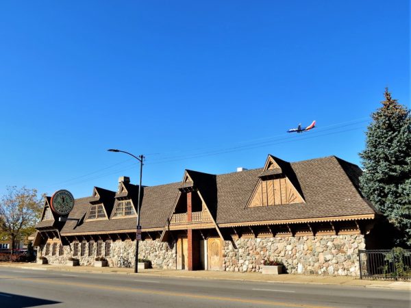 a tour bike leaning on the stone wall of a chalet style brown wood on story building with a plane in the air above.