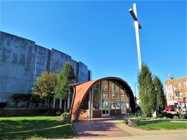 A tour bike at front of the Mid-century Modern glass brown metal saddle shaped entrance to an alternating stained glass and gray stone round church.