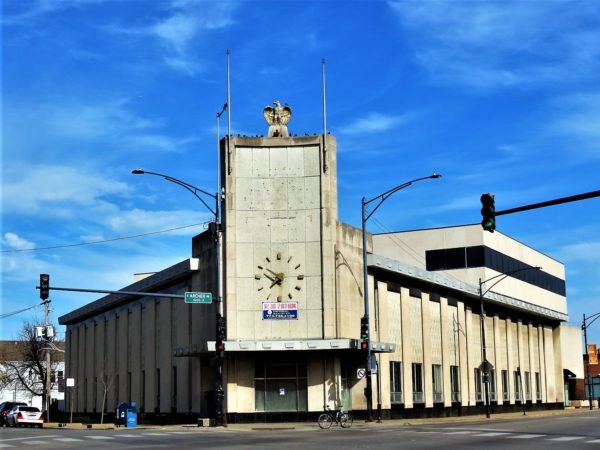 A tour bike at front of a concrete Midcentury two story diagonal corner former bank with a clock and eagle above the entry.