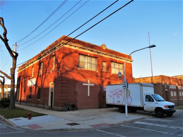 A tour bike leaning on a red brick corner building with two off color bricked up bay doors with a cross and a white delivery truck parked in front.