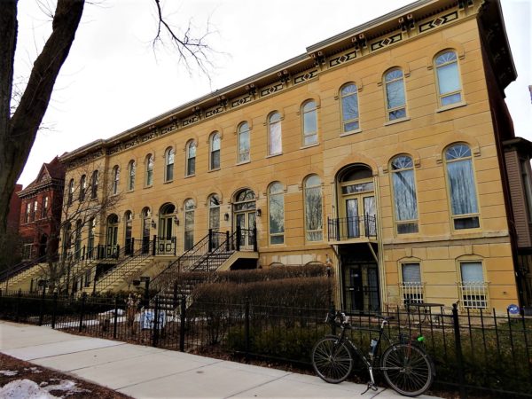 A tour bike leaning on a black metal fence in front of a three story yellow Italianate four section row house with stairs to second floor entrances.