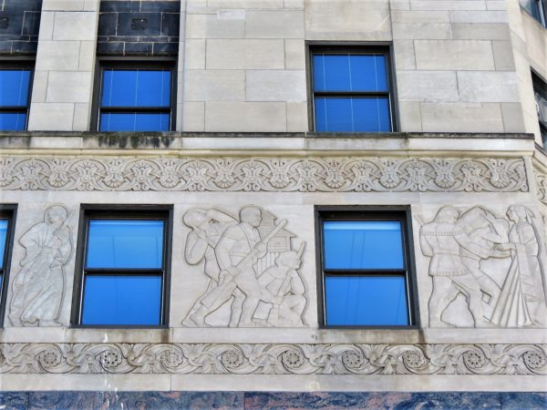 A limestone bas relief of Europeans and wood fort framed top and bottom by two design reliefs.