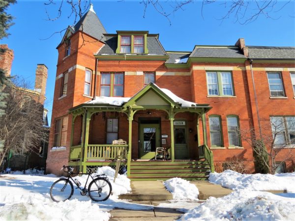 A tour bike standing in the snow in front of a red brick late 19th centruy home with a square corner turret and green painted wood front porch.