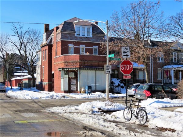 A tour bike standing across the street from a one story brick corner shop attached to a 19th century three story home.