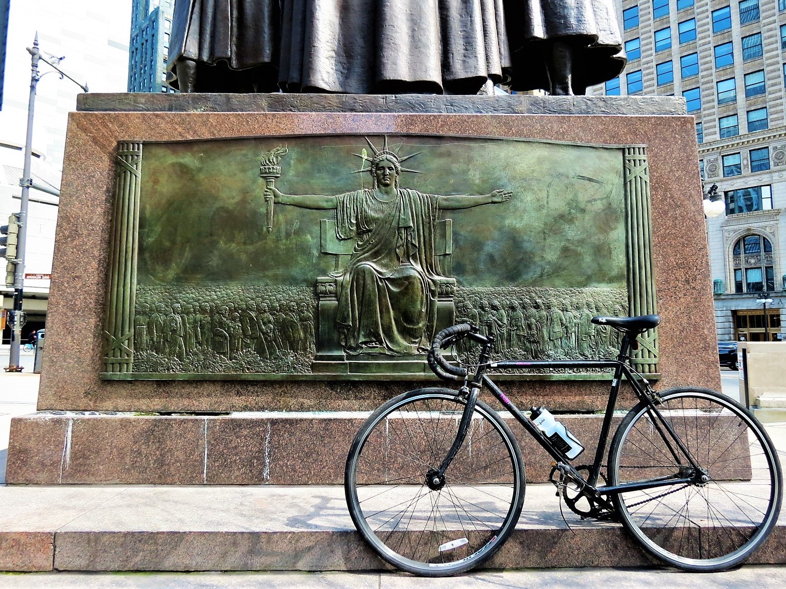 A tour bike leaning on a granite statue base embedded with a copper bas relief of Lady Liberty stretching her arms out over a crowd of people.