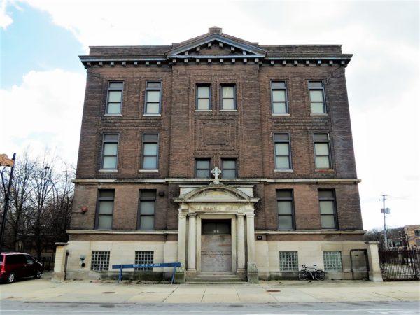 A tour bike leaning on the limestone first floor of a four story brown brick square mass school building with a cornice and pediment.