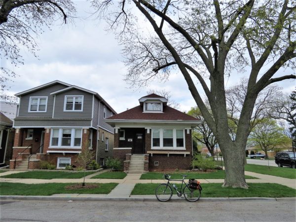 A tour bike at front of a one story brown brick raised bungalow with single central dormer.