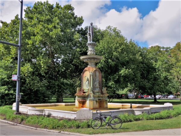 A tour bike standing at front of a patina bronze fountain with a male figure at top and trees behind.