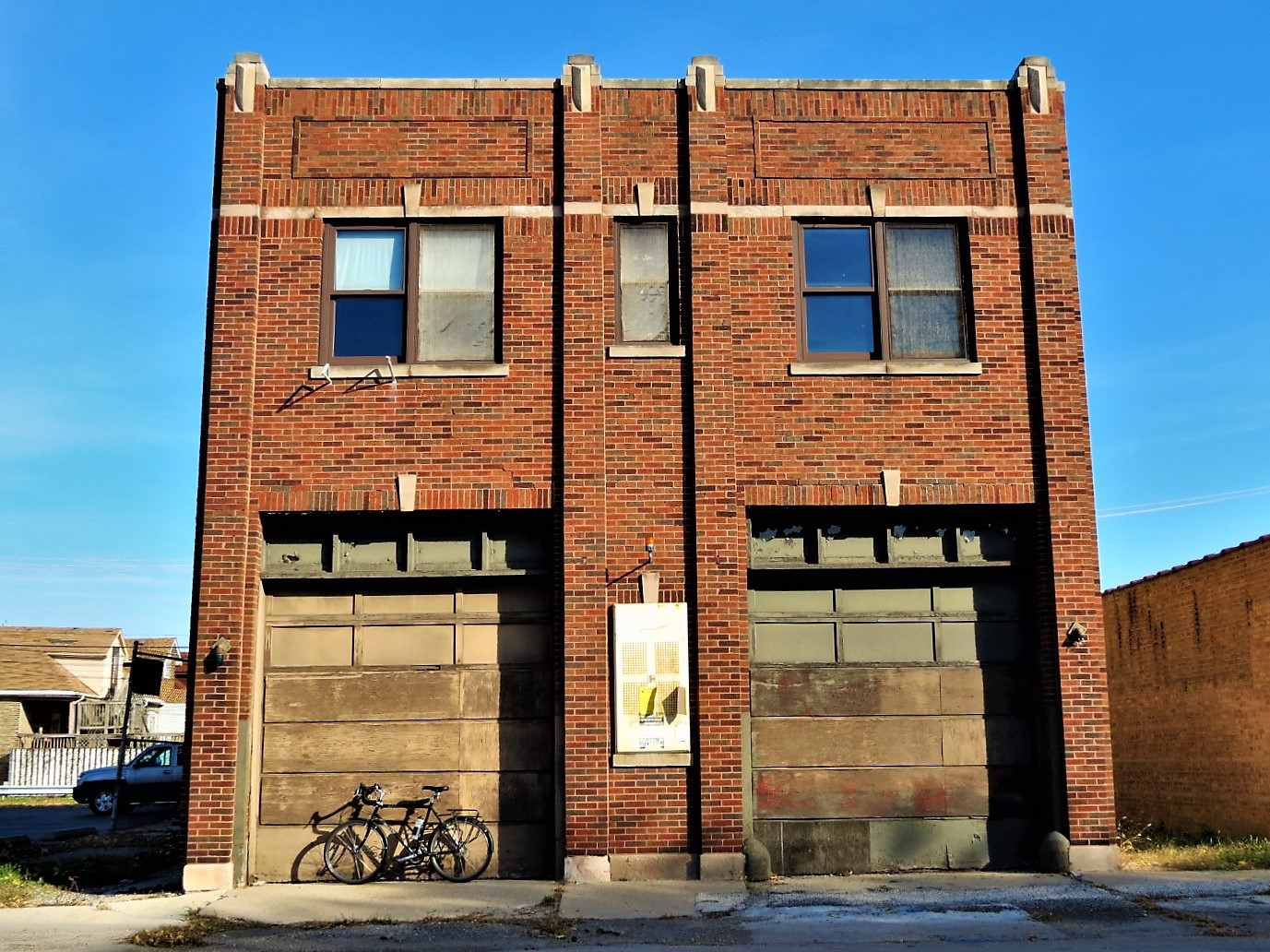 A tour bicycle leaning on the left of two high bay doors of a two story brown brick former firehouse.