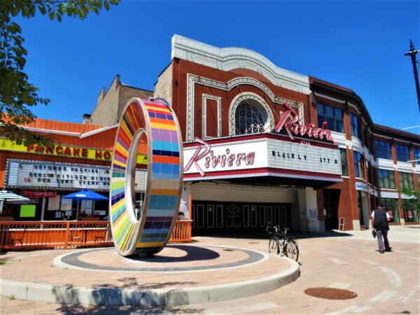 A tour bicycle standing between a rainbow colored ring and an early 19th centrury red brick and white terra cotta theatre with a white awning marquee.