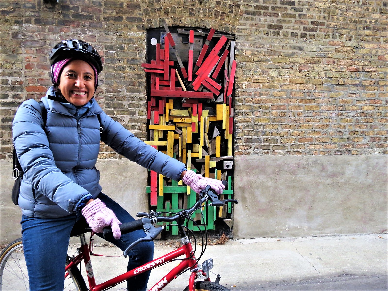 A helmeted CBA bike tour rider in front of an alley door covered in an abstract pattern of green, red, and yellow wooden slats smiling and looking at the camera.