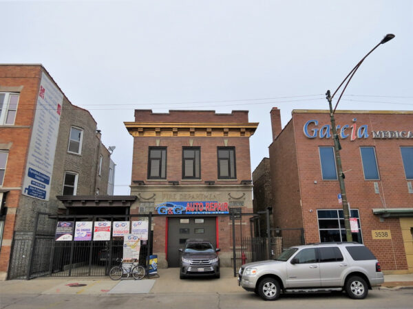 A tour bicycle leaning on the black metal fence in front of a two stroy brown brick former firehouse with a brown central bay door, yellow cornice, and auto parts sign.