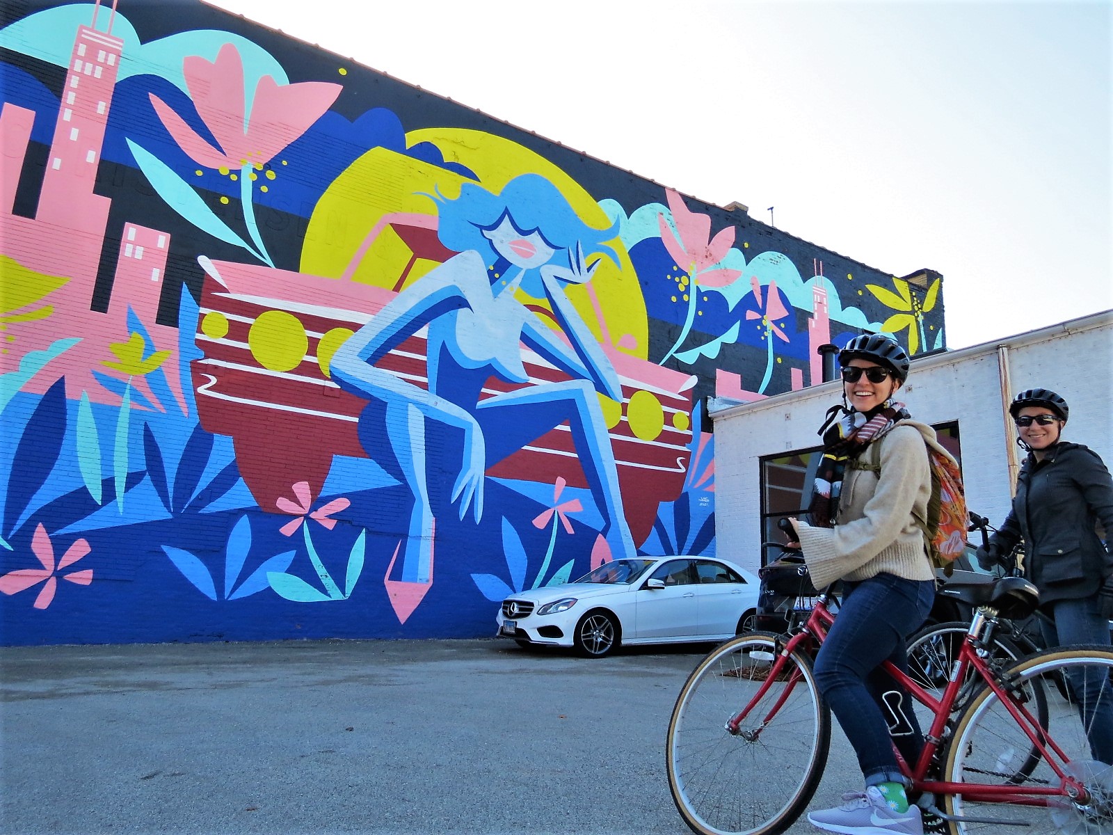 Two CBA bike tour riders smiling at the camera with brightly colored mural of a topless blue woman in high heels sitting in front pink Chicago buildings and a car.