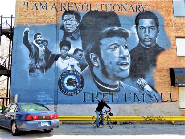 A CBA bike tour rider in front of a large black and white mural of Fred Hampton and other Black Panther figures.