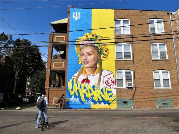 Two CBA bike tour riders looking a blue and yellow Ukraine and woman with flower head ring mural on an alley wall.