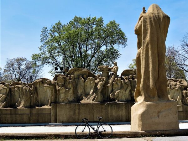 A tour bike leaning at front of the Fountain of Time, a large horizontal tan cement sculpture with one figure's back to the camera and many others facing front ensembled into a wave like formation.