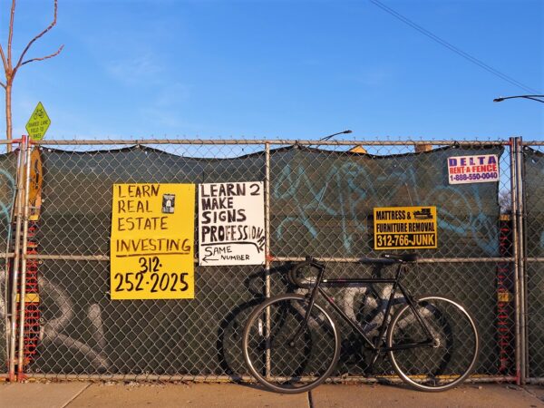 A tour bike leaning on a green clothed chain link fence with two hand written signs, one making fun of the other.