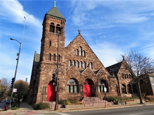 A tour bike at front of a corner late 1800s brown stone Richardsonian Romanesque church with bright red doors.
