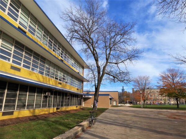 A tour bike standing to the side of a yellow brick three story mid-century modern school with a park in the near and the Willis Tower in the far distance.