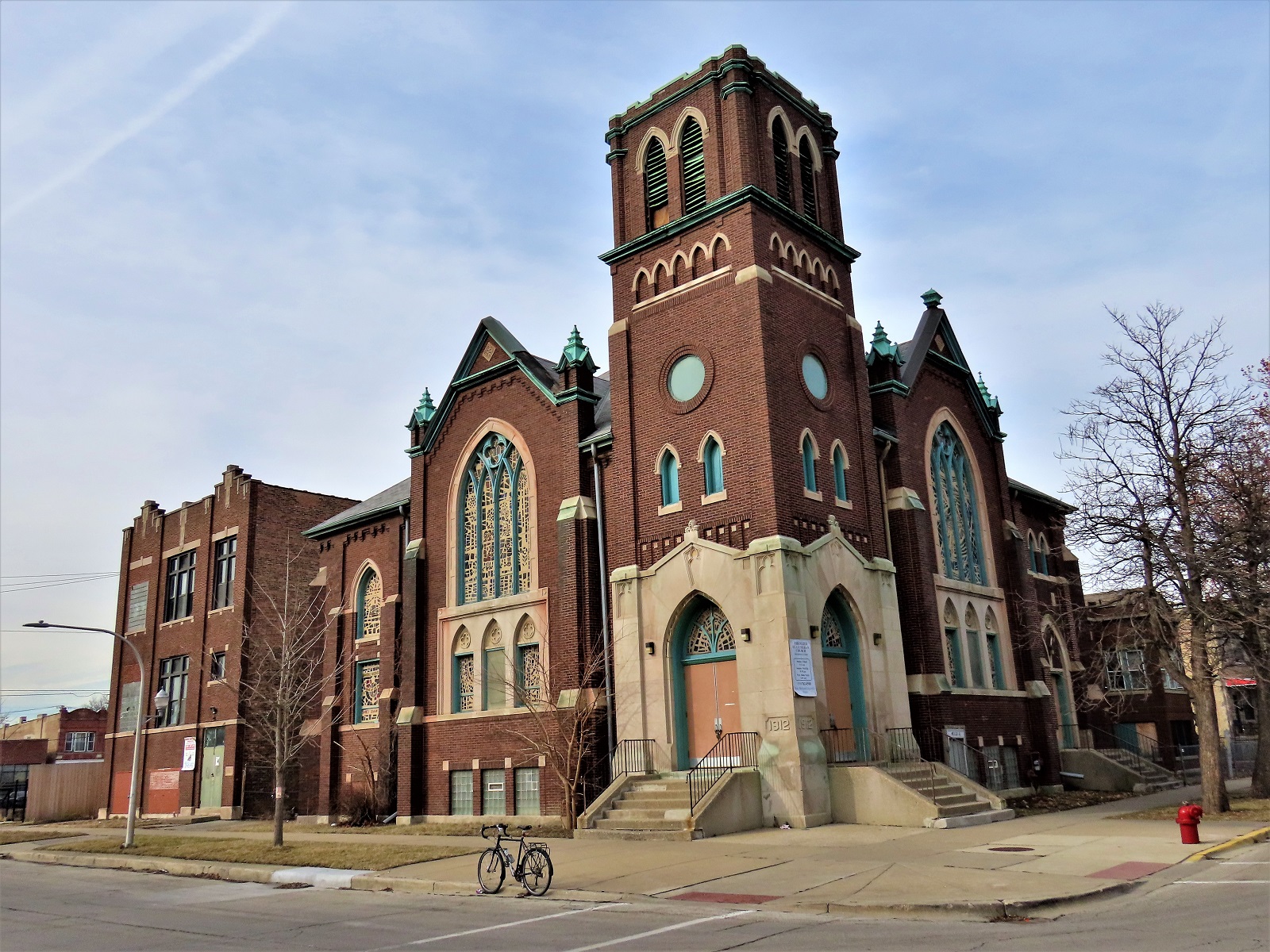 A tour bike standing at front of a brown brick, limestone detailed, pressed patina roofline double lot Gothic Revival corner church with school