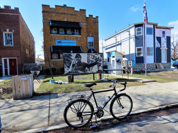 A tur bike at front of a tan brick two flat with a black and white poster of Fred Hampton in the front yard. panther prints on the sidewalk and free food box near the front door.