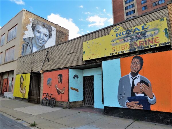 A tour bike leaning on a storefront boarded up with color blocked paintings that may reflect scenes from Lorraine Hansberry's Raisin in the Sun.