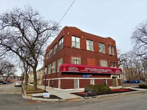 A tour bike standing at front of a three story red brick corner building with crosses of glass block windows and a new building length awning.