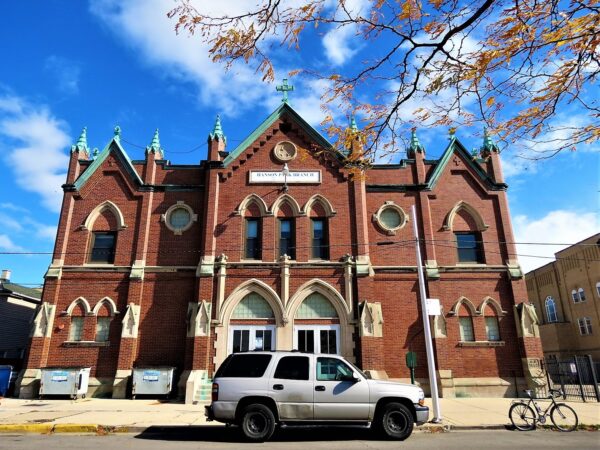 A tour bike standing with a white SUV at front of a two story double lot brown brick Gothic Revival church and school with copper roofline details.