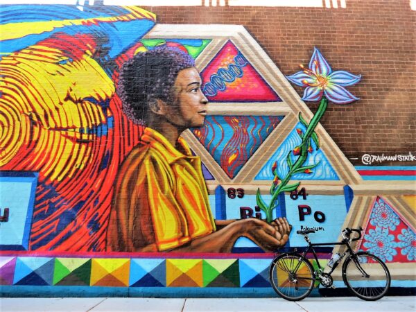 A tour bike leaning on a mural wall of an elder's face done in yellow behind a Black youth looking upward and holding a flower in his palm.