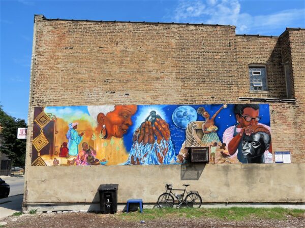A tour bike leaning on the common brick side of a building under a five segment horizontal mural depicting Black woman past and future.