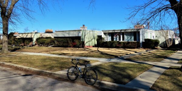 A tour bike standing at the curb in front of three windowed front wall midcentury modern box homes with medium plain lawn setbacks.