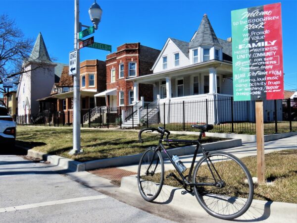 A tour bike standing below a green, black, and red vertical wooden sign that lays out what the block stands for with a wood Queen Anne home and brick two flats behind.