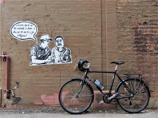 A tour bike leaning on a brown painted wall next to a wheatpaste of one man talking to another with a speech bubble.