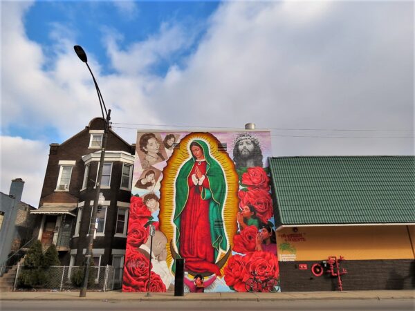 A tour bike leaning next to a three story mural of the Virgen de Guadeloupe surrounded by sepia colored faces of Jesus Christ on the right and three version of the same woman on the left.