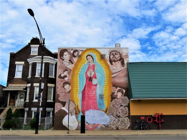 A tour bike leaning next to a three story mural of the Virgen de Guadeloupe surrounded by sepia colored faces of Jesus Christ and other people.