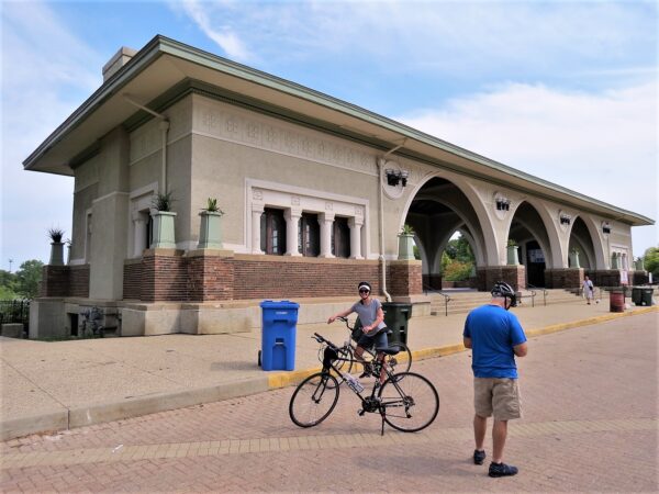 Two CBA tour riders in front of a wide Prairie School style boathouse with a low hipped roof and three middle arches.