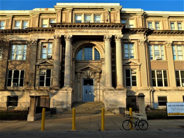 A tour bike standing at the entry steps of a limestone three story Classical Revival high school building bathed in evening sun.