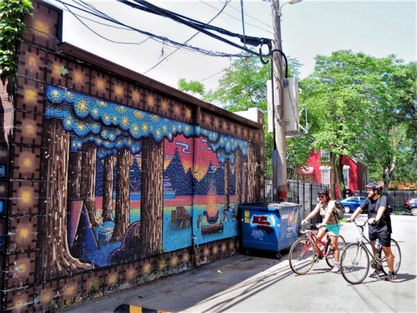 Two CBA tour riders looking at an alley mural of campfire in a forest made from colored dots.