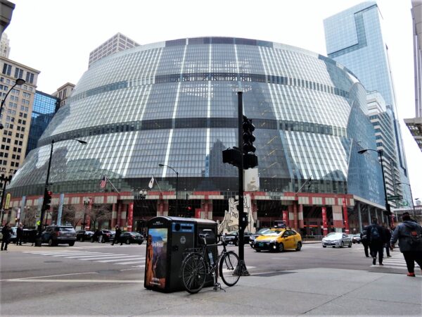 A tour bike leaning on a black trash box with the four story round glass atrium of the Thompson Center behind.