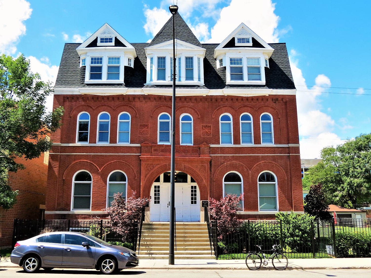 A tour bike standing at front of a three story double lot Queen Anne red brick hall with an arched center entrance