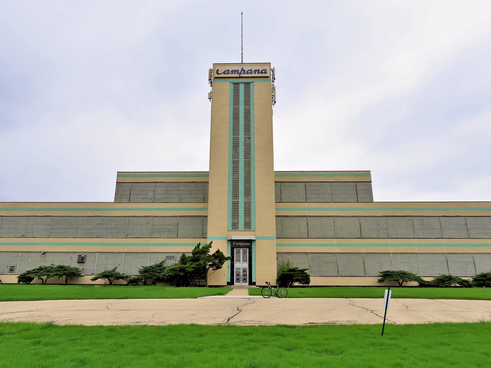 A tour bike standing in front of a massive yellow and green accent brick Streamline Moderne three story factory with five story central entryway tower.