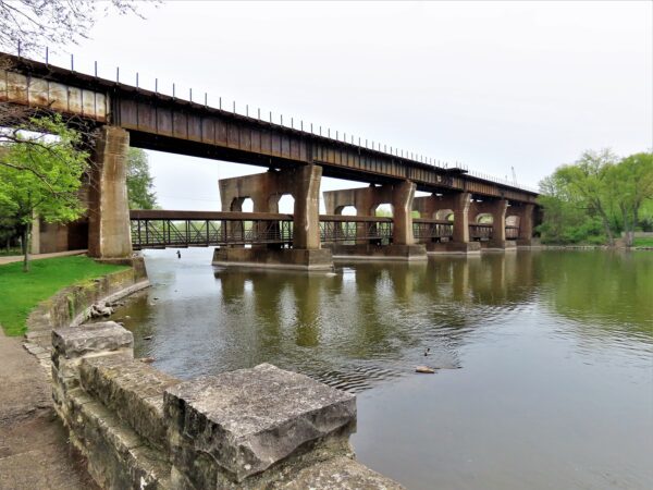 A barely visible tour bike on a pedestrian bridge running underneath a rail road bridge over a calm flowing river, a fisher visible in the distance.