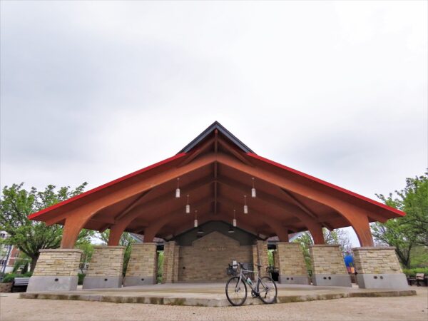 A tour bike standing under a Japanese and Prairie School inspired orange wood band shell on thin rock pillars. 