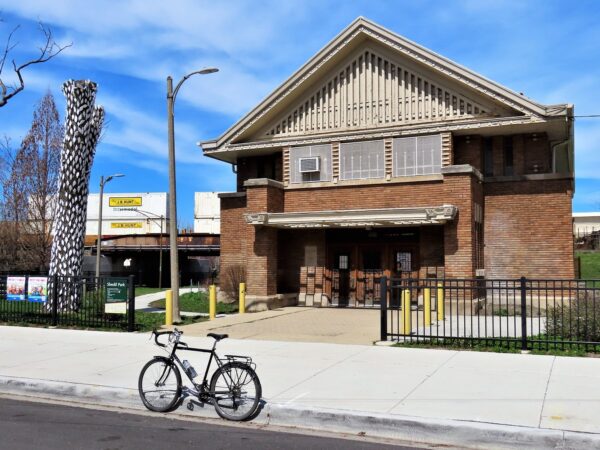 A tour bike standing at front of a tan brick Prairie School park building with a white scaled tree sculpture to the left and freight train behind.