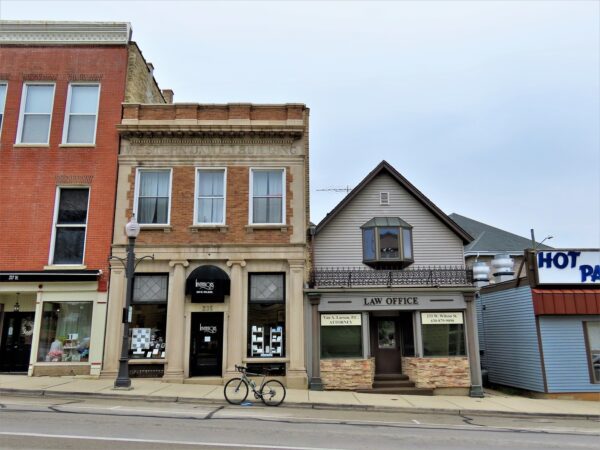 A tour bike standing at front of a two story brick storefreont and flat with Western United Building epigraph amd a one and half story wood storefront and attic sitting on a sloped street.