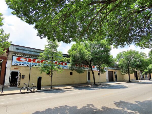 A bicycle standing at front of a one story green & white glazed brick building with yellow bricked yo storefronts, with a green leaf canopy.