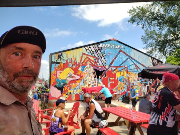 Me facing the camera with cyclist sitting at red picnic tables and a beer themed mural behind.