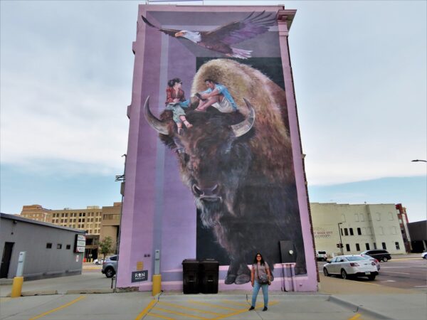 A woman sanding in front of a four story purple based mural with two people sitting on the head of a bison with an eagle lying over head.