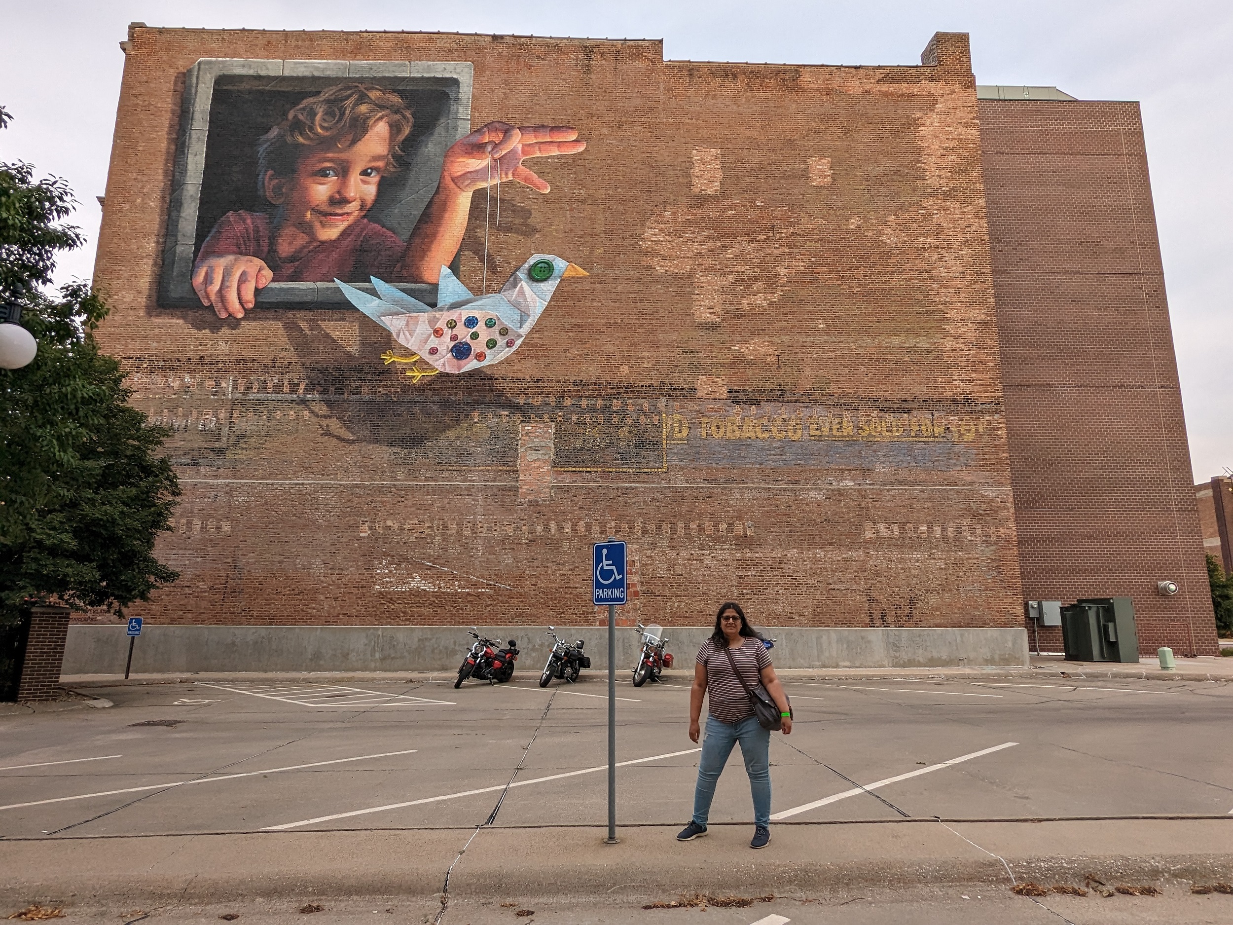 A woman standing in front of a three story brick side wall with a tobacco ghost sign and a mural of a boy with a paper and button bird puppet.